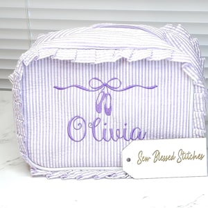 May include: A lavender and white striped cosmetic bag with a ruffled edge. The bag features embroidered ballet shoes and the name "Olivia" in lavender script. A tag with the text "Sew Blessed Stitches" is attached.