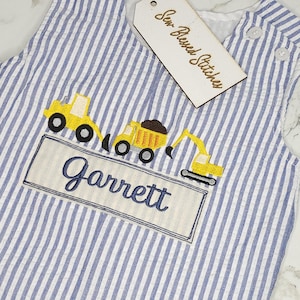 Peut inclure: Une salopette en seersucker à rayures bleues et blanches avec une broderie de véhicules de chantier jaunes et le prénom Garrett en écriture bleue foncée. Une étiquette blanche avec le texte Sew Blessed Stitches est fixée au vêtement.