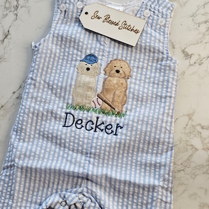 Pu&ograve; includere: Una tutina in seersucker azzurro e bianco con un ricamo di due cani, uno con un berretto blu e l'altro con una mazza da baseball e una palla. Il nome Decker &egrave; ricamato in blu sotto i cani. Un'etichetta riporta la scritta Sew Blessed Stitches.