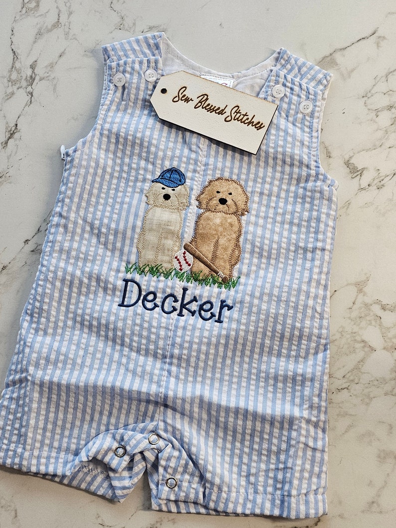 Pu&ograve; includere: Una tutina in seersucker azzurro e bianco con un ricamo di due cani, uno con un berretto blu e l'altro con una mazza da baseball e una palla. Il nome Decker &egrave; ricamato in blu sotto i cani. Un'etichetta riporta la scritta Sew Blessed Stitches.