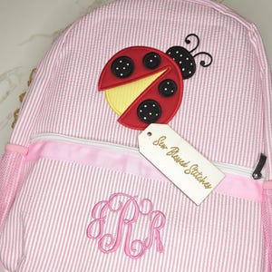 May include: A pink and white seersucker backpack featuring a red ladybug appliqué with black polka dots and a yellow accent. The front pocket displays a pink embroidered monogram with the letters RRR and a tag labeled Sew Blessed Stitches.