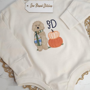 May include: An off-white long-sleeve bodysuit featuring an embroidered tan dog wearing a green, blue, and white plaid scarf, next to two stacked pumpkins and the navy blue letters "JD". A white tag with "Sew Blessed Stitches" is attached.