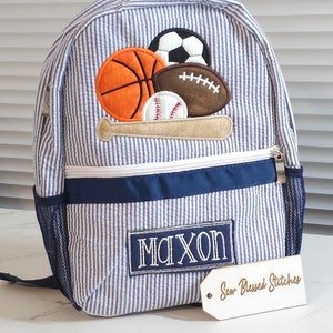 May include: Blue and white striped backpack with a navy blue trim and a mesh pocket. The backpack has an embroidered patch with a baseball bat and various sports balls, including a baseball, basketball, football, and soccer ball. The backpack also has a personalized name tag that says "MAXON" and a wooden tag that says "Sew Blessed Stitches".