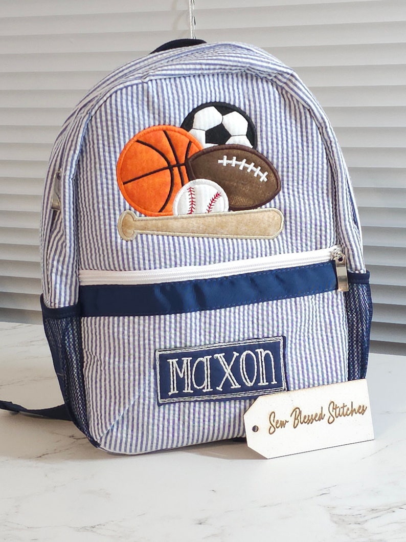 May include: Blue and white striped backpack with a navy blue trim and a mesh pocket. The backpack has an embroidered patch with a baseball bat and various sports balls, including a baseball, basketball, football, and soccer ball. The backpack also has a personalized name tag that says "MAXON" and a wooden tag that says "Sew Blessed Stitches".
