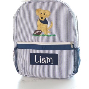Peut inclure: Sac &agrave; dos ray&eacute; bleu et blanc avec une applique de chien jaune et de ballon de football. Le sac &agrave; dos a une bordure bleu marine et une poche en filet sur le c&ocirc;t&eacute;. Le nom "Liam" est brod&eacute; sur le devant du sac &agrave; dos.
