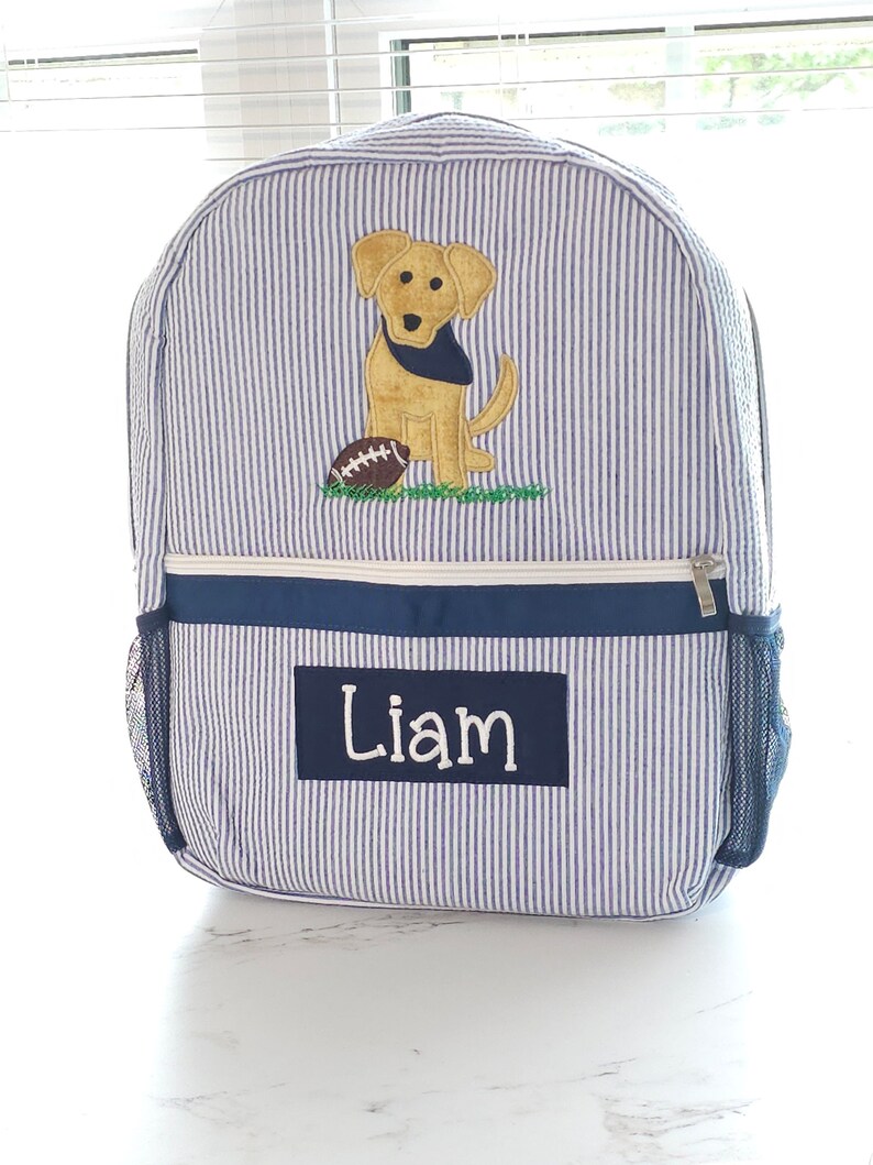 Peut inclure: Sac &agrave; dos ray&eacute; bleu et blanc avec une applique de chien jaune et de ballon de football. Le sac &agrave; dos a une bordure bleu marine et une poche en filet sur le c&ocirc;t&eacute;. Le nom "Liam" est brod&eacute; sur le devant du sac &agrave; dos.