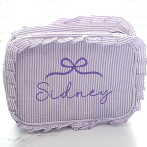 May include: A lavender and white striped bag with a ruffled edge. The bag features the name "Sidney" embroidered in a matching lavender color, along with a bow design above the name. The bag is rectangular in shape.