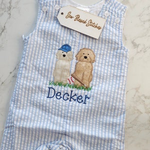 Pu&ograve; includere: Una tutina in seersucker azzurro e bianco con un ricamo raffigurante due cani, una palla da baseball e una mazza. Un cane indossa un berretto blu. Il nome Decker &egrave; ricamato in filo blu sotto i cani. Un'etichetta riporta Sew Blessed Stitches.