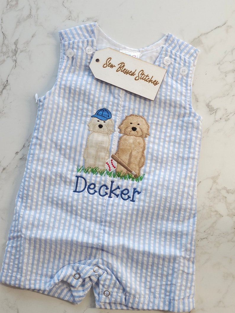 Pu&ograve; includere: Una tutina in seersucker azzurro e bianco con un ricamo raffigurante due cani, una palla da baseball e una mazza. Un cane indossa un berretto blu. Il nome Decker &egrave; ricamato in filo blu sotto i cani. Un'etichetta riporta Sew Blessed Stitches.