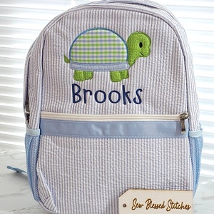 May include: A blue and white striped backpack with a green turtle applique and the name "Brooks" embroidered on the front. The backpack has a blue zipper and a blue and white striped handle. A wooden tag with the text "Sew Blessed Stitches" is in the foreground.