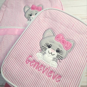 May include: Pink and white striped backpack with a gray cat with a pink bow embroidered on it. The backpack has a pink strap and the name Genevieve is embroidered on the front.