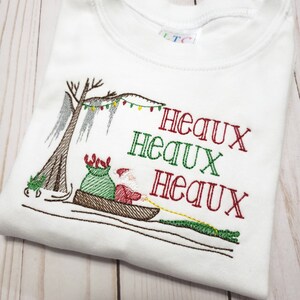 May include: A white t-shirt featuring an embroidered design of Santa Claus in a pirogue boat with a green sack, pulled by an alligator. A cypress tree with string lights hangs above. The text "HEAUX HEAUX HEAUX" is embroidered in red and green.