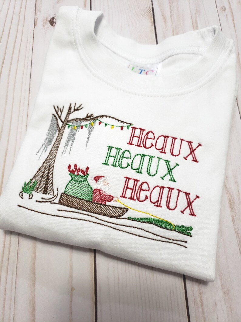 May include: A white t-shirt featuring an embroidered design of Santa Claus in a pirogue boat with a green sack, pulled by an alligator. A cypress tree with string lights hangs above. The text "HEAUX HEAUX HEAUX" is embroidered in red and green.