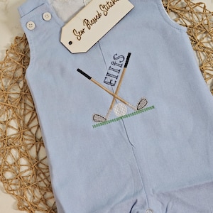 May include: Pale blue baby romper featuring embroidered golf clubs, a golf ball, and the name "Ellis". The romper has white buttons and a tag that reads "Sew Blessed Stitches".