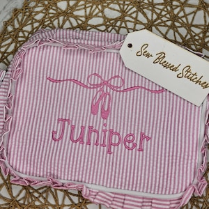 May include: A pink and white striped bag with a ruffled edge. The bag features an embroidered design of ballet shoes and the name "Juniper" in pink. A tag with the text "Sew Blessed Stitches" is attached.