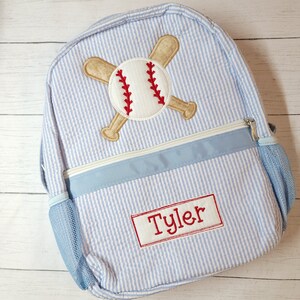 K&ouml;nnte beinhalten: Ein blauer und wei&szlig; gestreifter Rucksack mit einem Baseball- und gekreuzten Fledermaus-Design. Der Rucksack hat einen blauen Rei&szlig;verschluss und eine Netztasche an der Seite. Der Name "Tyler" ist auf der Vorderseite des Rucksacks gestickt.