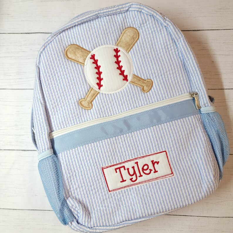 K&ouml;nnte beinhalten: Ein blauer und wei&szlig; gestreifter Rucksack mit einem Baseball- und gekreuzten Fledermaus-Design. Der Rucksack hat einen blauen Rei&szlig;verschluss und eine Netztasche an der Seite. Der Name "Tyler" ist auf der Vorderseite des Rucksacks gestickt.