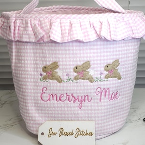 May include: Pink and white gingham Easter basket with a ruffled top and handle. The basket features embroidered brown bunnies and the name "Emersyn Mae" in pink script. A wooden tag reads "Sew Blessed Stitches."