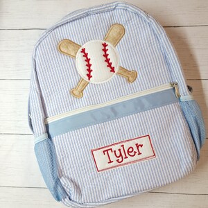 K&ouml;nnte beinhalten: Ein blauer und wei&szlig; gestreifter Rucksack mit einem Baseball- und Schl&auml;ger-Design. Der Rucksack hat eine blaue Netztasche an der Seite und eine blaue Rei&szlig;verschlusstasche vorne. Der Name "Tyler" ist auf der Vorderseite des Rucksacks gestickt.