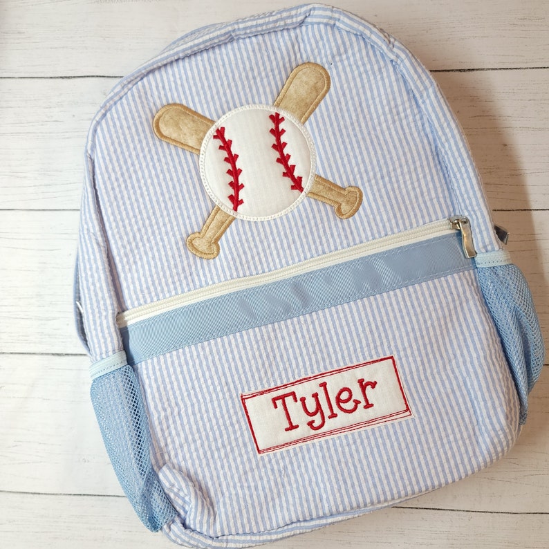 K&ouml;nnte beinhalten: Ein blauer und wei&szlig; gestreifter Rucksack mit einem Baseball- und Schl&auml;ger-Design. Der Rucksack hat eine blaue Netztasche an der Seite und eine blaue Rei&szlig;verschlusstasche vorne. Der Name "Tyler" ist auf der Vorderseite des Rucksacks gestickt.