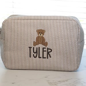 Könnte beinhalten: Eine rechteckige Kosmetiktasche mit Seersucker-Streifen in Beige und Weiß. Die Tasche hat einen gestickten Teddybären und den Namen "TYLER" in braunem Faden. Die Tasche hat einen Reißverschluss und abgerundete Ecken.