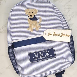 May include: Blue and white striped backpack with a tan dog applique wearing a blue bandana. The backpack has a navy blue trim, mesh side pocket, and the name "JACK" embroidered on a navy blue patch. A tag reads "Sew Blessed Stitches".