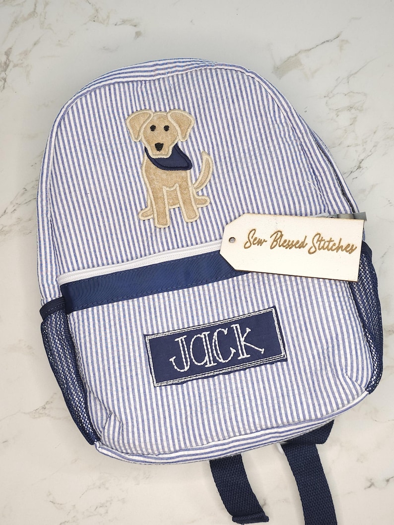 May include: Blue and white striped backpack with a tan dog applique wearing a blue bandana. The backpack has a navy blue trim, mesh side pocket, and the name "JACK" embroidered on a navy blue patch. A tag reads "Sew Blessed Stitches".