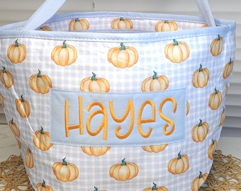 May include: A fabric basket with a light blue handle and a pattern of orange pumpkins on a blue and white gingham background. The name "Hayes" is embroidered in orange on a blue patch. The basket is on a woven mat.