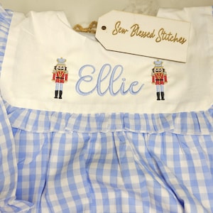 May include: A blue and white gingham patterned garment with a white yoke featuring embroidered nutcracker soldiers and the name "Ellie" in blue script. A tag with "Sew Blessed Stitches" is attached.