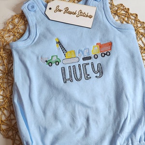 May include: Light blue baby onesie with an embroidered design of construction vehicles and the name "HUEY".