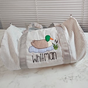 May include: A striped duffel bag with gray and white vertical stripes. The bag features an embroidered duck design with the name "Whitman" in black lettering. The duck has a brown checkered body, green head, and orange beak.