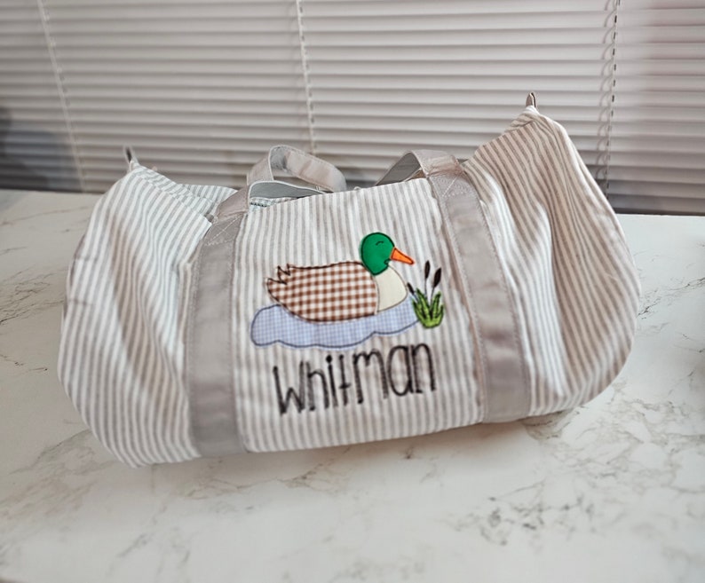 May include: A striped duffel bag with gray and white vertical stripes. The bag features an embroidered duck design with the name "Whitman" in black lettering. The duck has a brown checkered body, green head, and orange beak.
