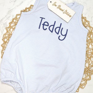 May include: Light blue and white striped baby romper with the name "Teddy" embroidered in navy blue. The romper has a round neckline and snap closures at the bottom. A wooden tag with the text "Sew Blessed Stitches" is attached.