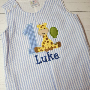 Peut inclure: Une grenouillère rayée bleu et blanche avec une girafe tenant un ballon et le chiffre un brodé sur le devant. Le prénom Luke est également brodé sur la grenouillère.