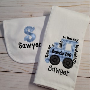 May include: A white burp cloth with blue and white checkered train embroidery. The train has the text "Train up a child in the way he should go" and "Proverbs 22:6". The burp cloth is personalized with the name "Sawyer".