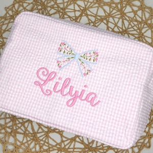 May include: Pink and white gingham cosmetic bag with the name "Lilyia" embroidered in pink script. A floral bow with pink and blue accents is embroidered above the name. The bag is rectangular and has a zipper closure.