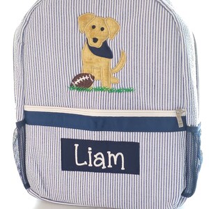 Peut inclure: Sac &agrave; dos ray&eacute; bleu et blanc avec un chien jaune et un ballon de football brod&eacute;s sur le devant. Le sac &agrave; dos a une poche zipp&eacute;e bleu marine et une poche lat&eacute;rale en filet. Le nom "Liam" est brod&eacute; sur la poche avant.