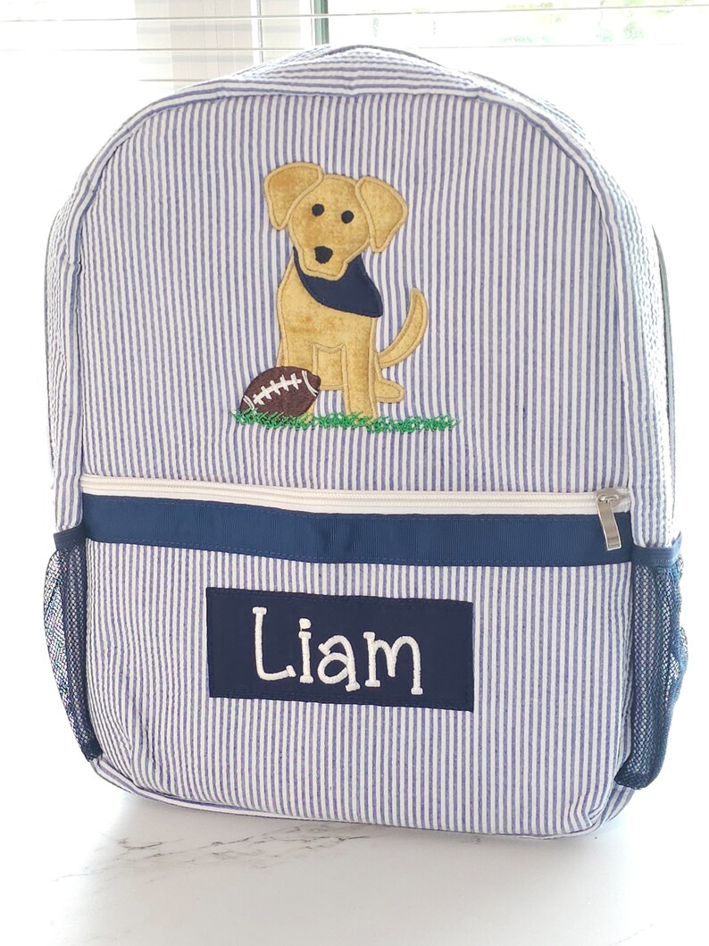 Peut inclure: Sac &agrave; dos ray&eacute; bleu et blanc avec un chien jaune et un ballon de football brod&eacute;s sur le devant. Le sac &agrave; dos a une poche zipp&eacute;e bleu marine et une poche lat&eacute;rale en filet. Le nom "Liam" est brod&eacute; sur la poche avant.