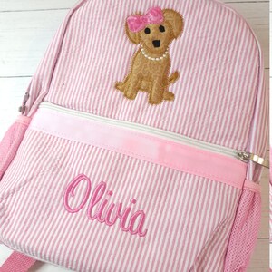 May include: Pink and white striped backpack with a brown dog embroidered on the front. The dog is wearing a pink bow and a pearl necklace. The name "Olivia" is embroidered on the front of the backpack.