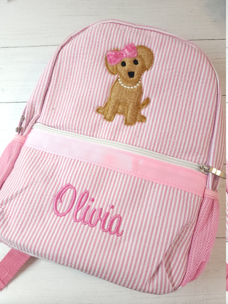 May include: Pink and white striped backpack with a brown dog embroidered on the front. The dog is wearing a pink bow and a pearl necklace. The name "Olivia" is embroidered on the front of the backpack.