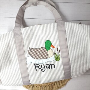 May include: A gray and white striped tote bag with a duck applique. The duck has a green head, brown and white checkered body, and an orange beak. The name "Ryan" is embroidered below the duck in black script. The bag has gray handles and trim.
