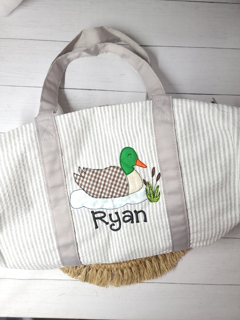 May include: A gray and white striped tote bag with a duck applique. The duck has a green head, brown and white checkered body, and an orange beak. The name "Ryan" is embroidered below the duck in black script. The bag has gray handles and trim.