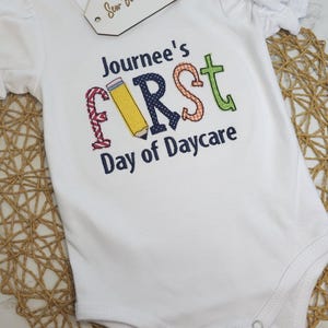 K&ouml;nnte beinhalten: Wei&szlig;er Strampler mit dem Text "Journee's First Day of Daycare" in bunten gestickten Buchstaben. Der Buchstabe "F" ist ein gelber Bleistift.