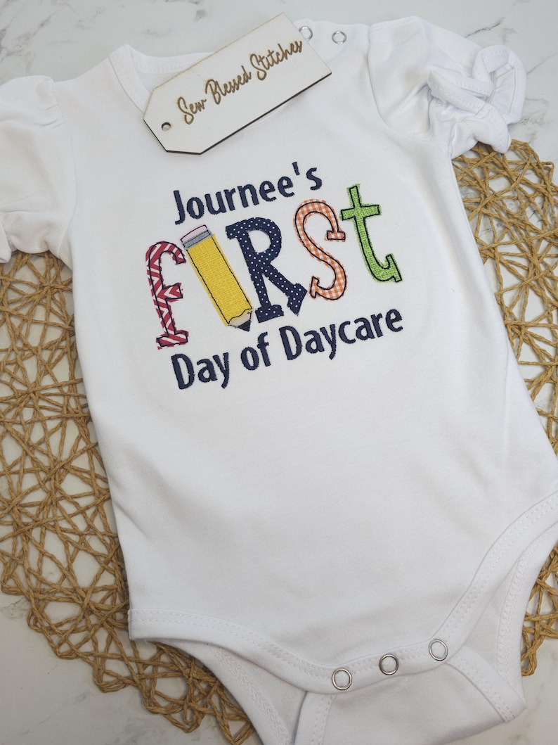 K&ouml;nnte beinhalten: Wei&szlig;er Strampler mit dem Text "Journee's First Day of Daycare" in bunten gestickten Buchstaben. Der Buchstabe "F" ist ein gelber Bleistift.