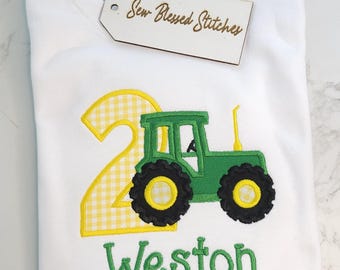 May include: A white short-sleeved t-shirt featuring an embroidered green tractor and a large yellow gingham number two. The name Weston is embroidered in green below the tractor. A wooden tag with the text Sew Blessed Stitches is attached.