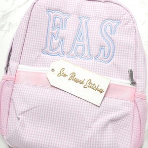 May include: A light pink and white gingham backpack featuring the embroidered letters EAS in light blue thread. The backpack includes a white zipper, a pink ribbon accent, and a white tag with the text Sew Blessed Stitches printed in gold.