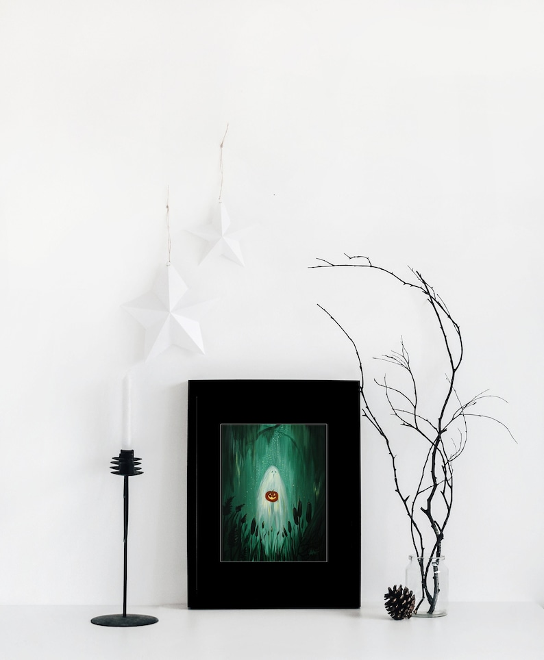 May include: A framed painting of a ghost holding a jack-o'-lantern in a dark green forest. The painting is in a black frame and is hanging on a white wall.