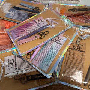Könnte beinhalten: Eine Sammlung verpackter Geschenke mit Stiften, Schlüsselanhängern und Lesezeichen. Jedes Paket enthält einen Stift, einen Schlüsselanhänger mit Lederriemen und ein Lesezeichen mit dem Text "Bible Emergency Numbers". Die Pakete sind in klarem Kunststoff versiegelt.