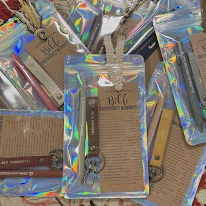 以下が含まれることがあります： 「Bible EMERGENCY NUMBERS」の文字が入ったペン、キーホルダー、タグが入った透明な虹色のポーチ。ポーチは模様のある表面に配置され、さまざまな色と質感があります。