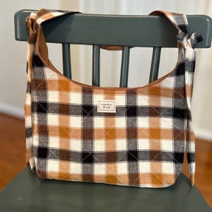 May include: A quilted hobo bag featuring a plaid pattern in shades of brown, black, and white. The bag has a wide shoulder strap with knotted ends and a rectangular fabric label centered on the front that reads VIRGINIA & RAE in black text.